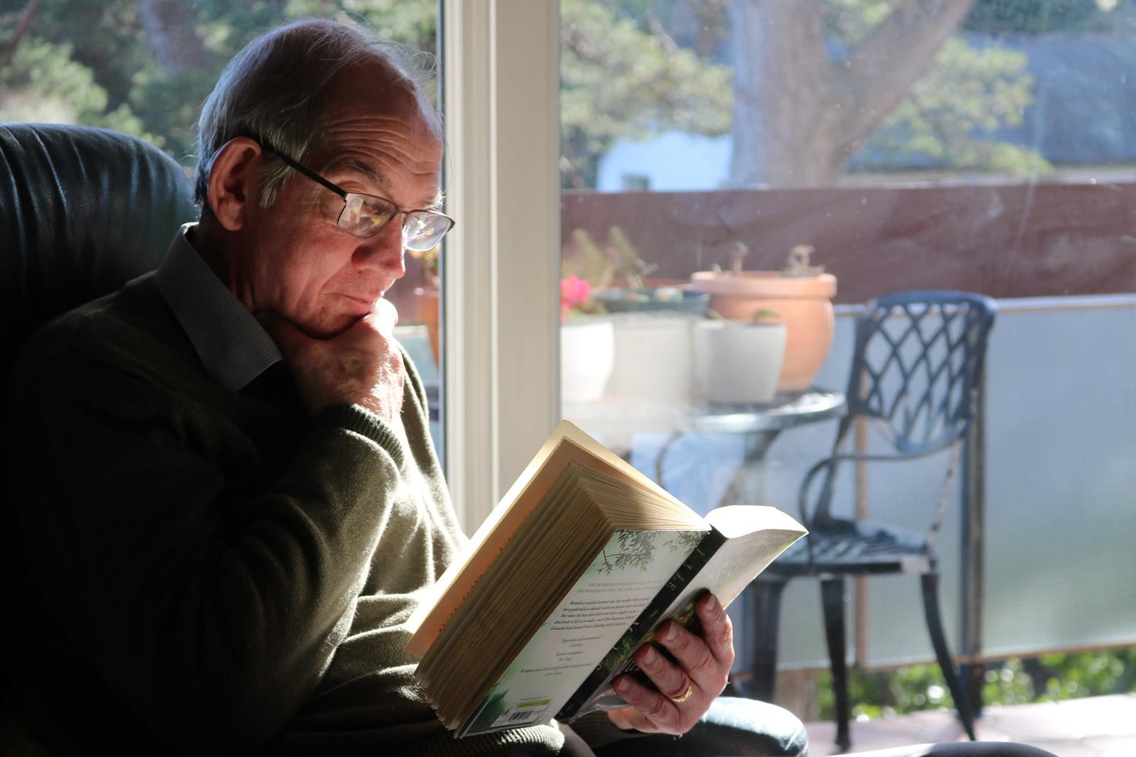 Um homem lê um livro perto de uma janela iluminada, com plantas ao fundo.